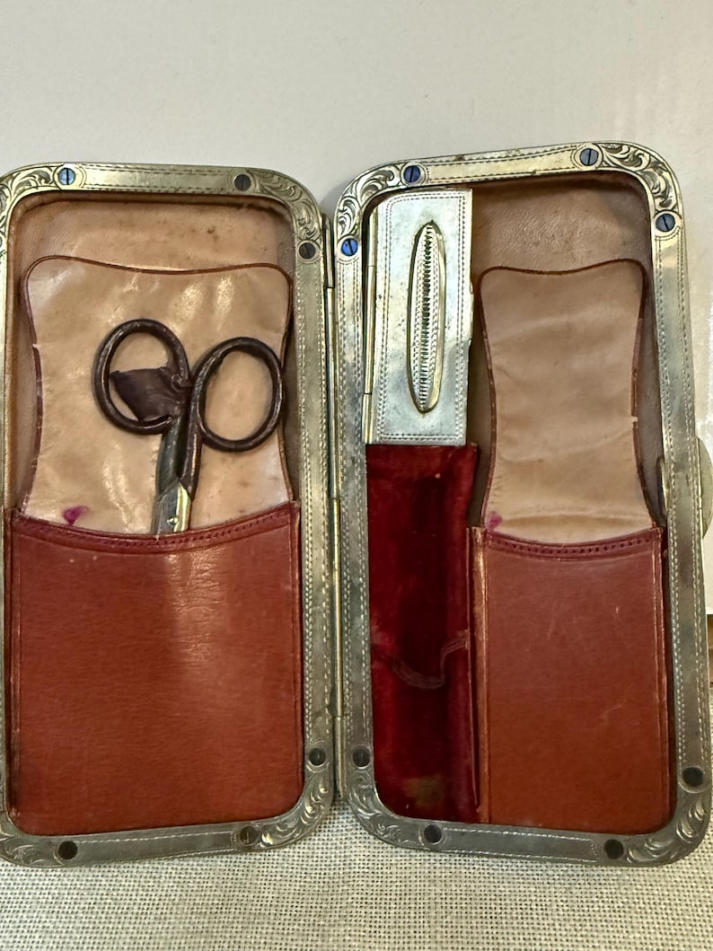 May include: Open antique travel grooming kit with a silver-toned frame and decorative edges. The interior features brown leather pockets, a pair of scissors, and a small silver case. The lining is tan leather and red velvet.