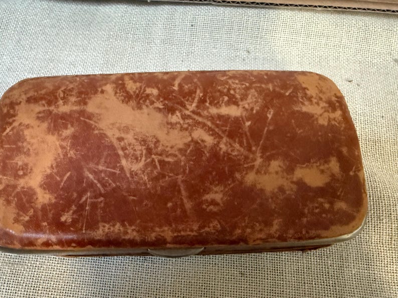 May include: A rectangular, vintage brown leather case with a distressed appearance. The case has a metal trim and a clasp closure. The leather shows signs of wear and tear, with lighter patches and scratches, giving it a rustic look.