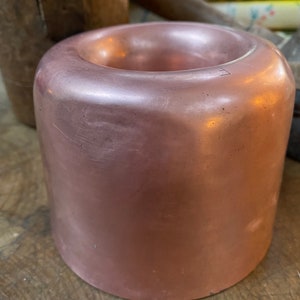 May include: A rose gold colored metal cup with a smooth, shiny finish. The cup has a rounded shape and a slightly flared rim.