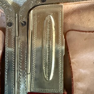May include: Close-up of a vintage silver-toned metal compact. The compact features ornate detailing and a rectangular shape with a hinged lid. The interior reveals a red velvet lining and a light pink leather-like material.