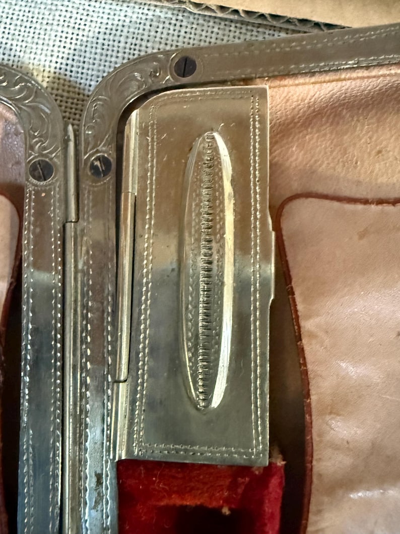 May include: Close-up of a vintage silver-toned metal compact. The compact features ornate detailing and a rectangular shape with a hinged lid. The interior reveals a red velvet lining and a light pink leather-like material.