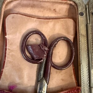May include: An antique pair of scissors with a dark brown handle and a silver blade, resting inside a leather case. The case is open, revealing a light tan interior and a burgundy leather exterior with a decorative metal frame.