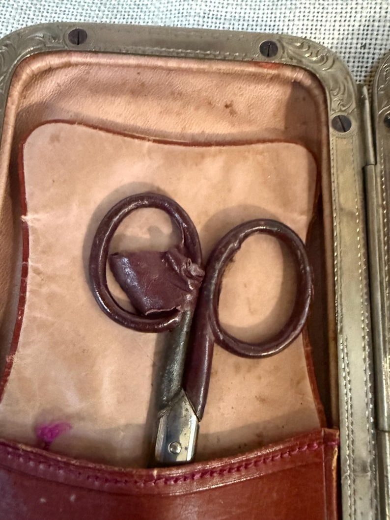 May include: An antique pair of scissors with a dark brown handle and a silver blade, resting inside a leather case. The case is open, revealing a light tan interior and a burgundy leather exterior with a decorative metal frame.