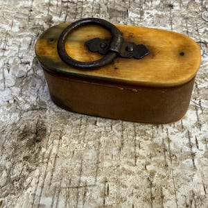 May include: An oval-shaped box with a light tan, smooth lid featuring a dark, weathered metal ring handle attached by a decorative metal plate. The sides of the box are a solid, matte brown color, creating a contrast with the lighter top.