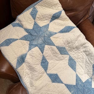 May include: A quilted blanket featuring a repeating star pattern in light blue and off-white. The fabric has a subtle, textured print within the blue sections, and the entire piece is finished with detailed diamond-shaped machine stitching.