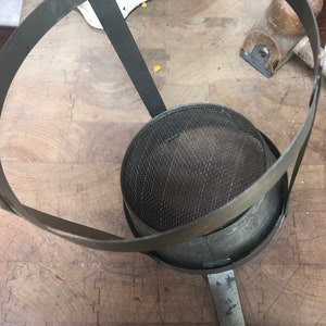 May include: A vintage metal strainer with a wire mesh bottom and a metal ring handle. The strainer is sitting on a wooden surface.