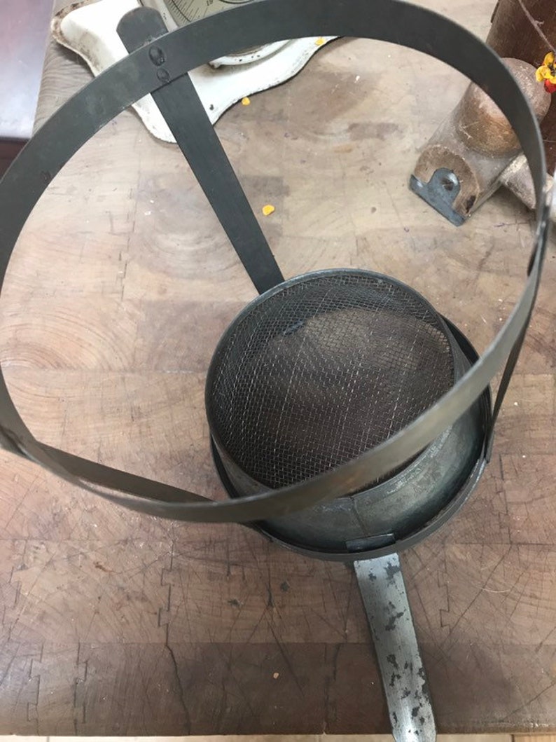 May include: A vintage metal strainer with a wire mesh bottom and a metal ring handle. The strainer is sitting on a wooden surface.