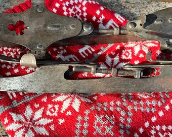 May include: Vintage ice skates with metal blades and a silver finish. The skates are resting on a red and white patterned fabric. The blades have a curved design, and the metal components show signs of wear.