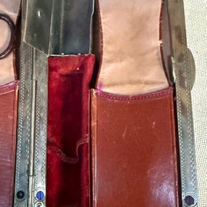 May include: Close-up of an antique leather and metal case. The case is open, revealing compartments lined with red velvet and tan leather. The metal frame has ornate detailing and blue and black accents.