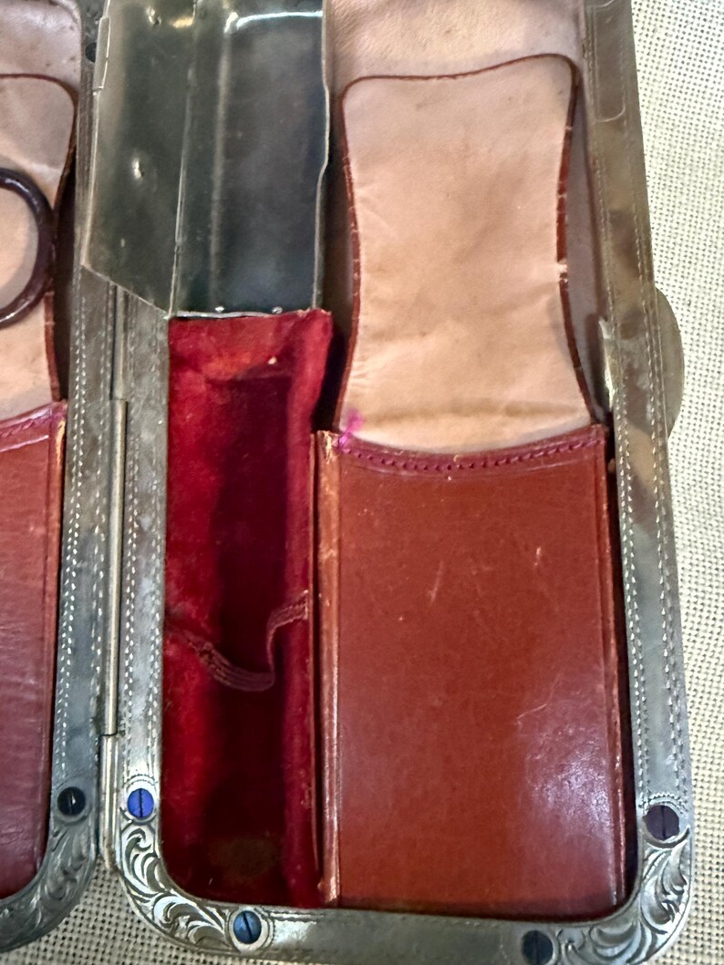 May include: Close-up of an antique leather and metal case. The case is open, revealing compartments lined with red velvet and tan leather. The metal frame has ornate detailing and blue and black accents.