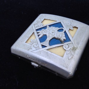May include: Silver-toned compact with floral design and the text "Cara Nome". The square compact features a blue and cream-colored center with an ornate floral motif. The edges are decorated with Art Deco style flourishes.