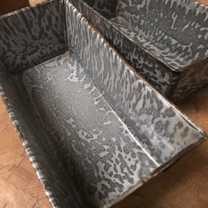 May include: Two vintage gray and white speckled metal loaf pans. The pans are nested together and have a rustic look.