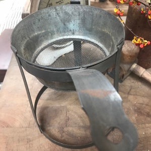 May include: A vintage metal strainer with a wire mesh bottom and a handle. The strainer is on a metal stand with a circular base.