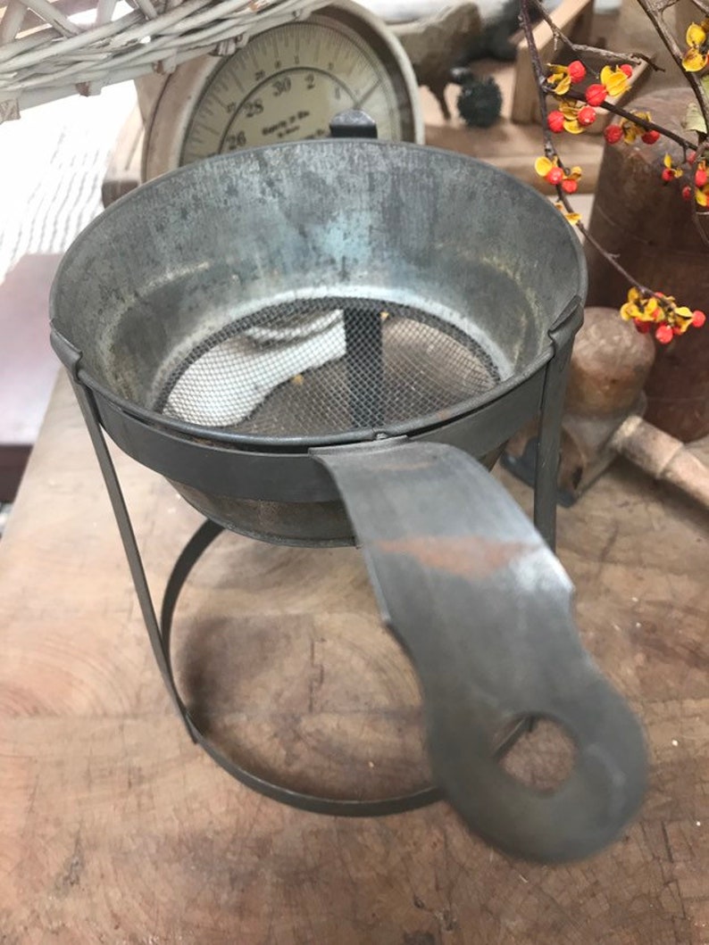May include: A vintage metal strainer with a wire mesh bottom and a handle. The strainer is on a metal stand with a circular base.