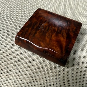 May include: A small, rectangular wooden box with a curved top. The box is made of a dark, reddish-brown wood with a swirling grain pattern. The box is closed and appears to be empty. The box is sitting on a light-colored, textured surface.