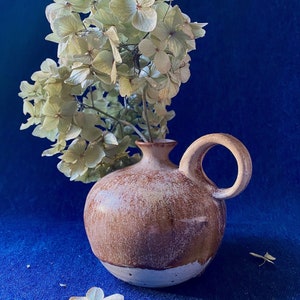 Può includere: Un piccolo vaso in ceramica marrone con manico, contenente ortensie essiccate. Il vaso ha una finitura maculata e una base bianca. Lo sfondo è un tessuto blu scuro, creando un contrasto.