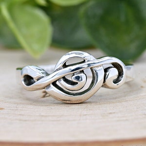 May include: A silver ring with a treble clef design. The ring is on a wooden surface.