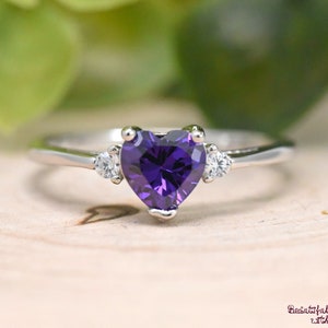 May include: A silver ring with a heart-shaped purple gemstone and two smaller clear gemstones on the band.