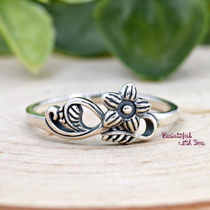May include: A silver ring with a delicate floral design. The ring features a stylized flower with a leaf on either side.