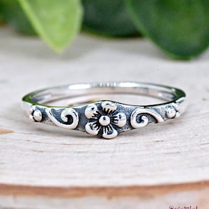 May include: A silver ring with a delicate floral design. The ring features a small flower with a swirl pattern on either side.