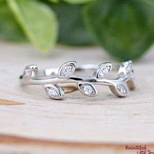 May include: A silver ring with a leaf design. The ring features small, round, clear gemstones set in the leaves. The ring is on a wooden surface with a blurred green background. The text "Beautiful with You" is in the bottom right corner.