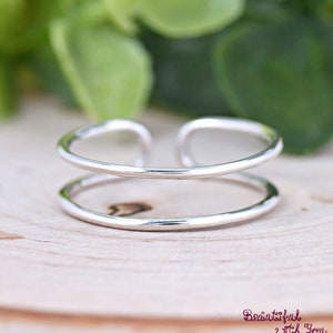 May include: A silver adjustable ring with a double band design. The ring is made of a smooth, polished metal and has a simple, minimalist style.