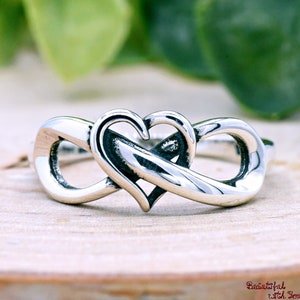 May include: A silver ring featuring a heart intertwined with an infinity symbol. The ring is made of polished metal and sits on a light-colored wooden surface. The background includes blurred green foliage.