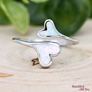 May include: A silver ring with two intertwined heart-shaped opal stones. The opals are iridescent and have a white base with pink and blue hues.