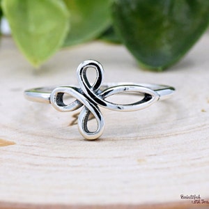 May include: A silver-colored ring with an openwork design. The ring features a central motif of intertwined loops, creating a stylized cross or floral shape. The band is thin and smooth, and the ring is set against a light-colored wooden surface.