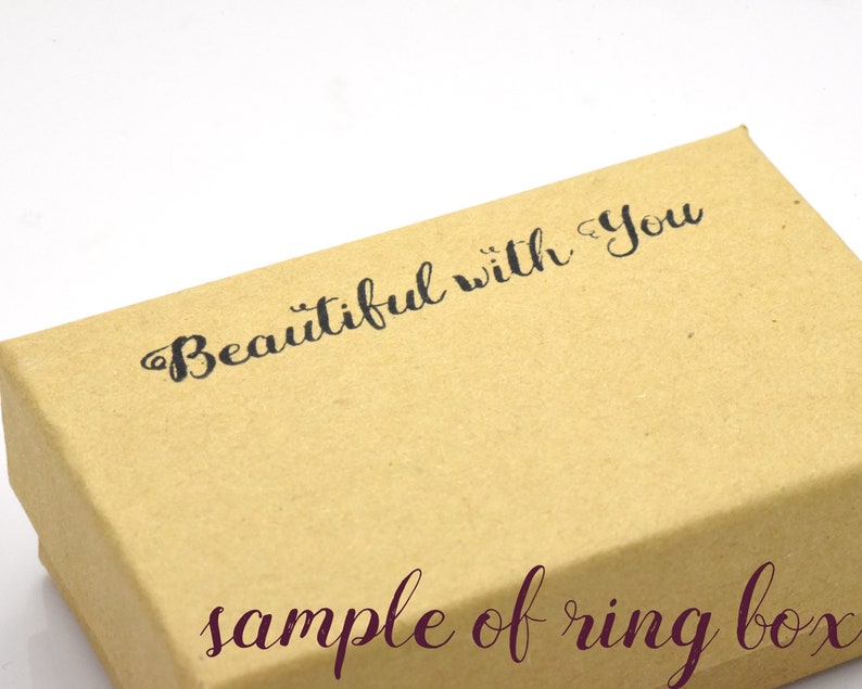 Puede incluir: Una caja de anillo de cart&oacute;n marr&oacute;n con el texto "Beautiful with you" impreso en la parte superior. Las palabras "sample of ring box" est&aacute;n impresas en la parte inferior de la caja en un tono marr&oacute;n m&aacute;s oscuro.