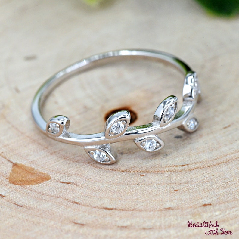 May include: A silver-colored ring with a leaf design. The ring features small, clear gemstones set along the leaves. The ring is displayed on a light-colored wooden surface. The text "Beautiful with You" is in the bottom right corner.