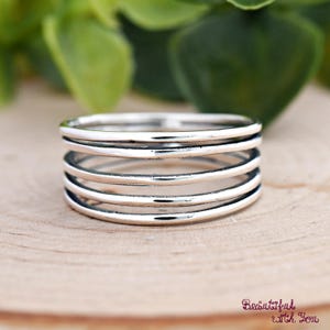 May include: A silver ring with five stacked bands. The ring is made of shiny metal and sits on a wooden surface. The background features green foliage. The text "Beautiful with You" is in the bottom right corner.