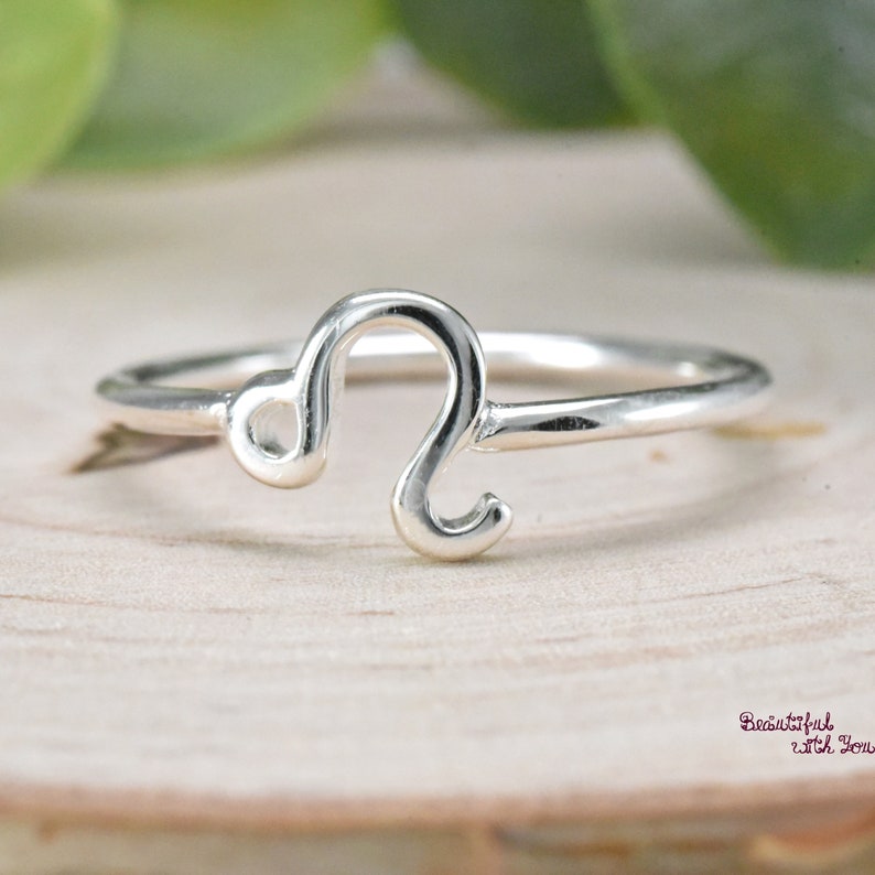 May include: A silver ring with a Leo zodiac symbol. The ring is thin and delicate, and the symbol is simple and elegant.
