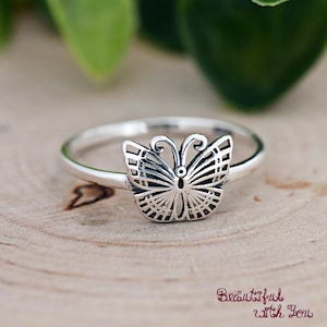 May include: A silver butterfly ring. The ring features a detailed butterfly design with openwork wings. The ring is set on a wooden surface with the text "Beautiful with You" in a cursive font.
