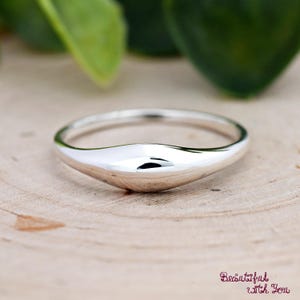 May include: A polished silver ring with a smooth, curved design. The ring has a sleek, modern aesthetic, reflecting light and creating a bright, reflective surface. The ring is set against a natural wood background.