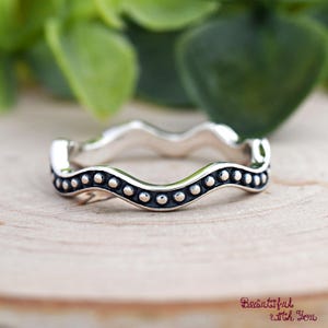 May include: A silver ring with a wavy design, accented with small, evenly spaced dots along the edges. The ring is set against a wooden surface with a blurred green background, creating a natural aesthetic. The ring's design is simple yet elegant.