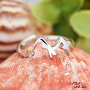 May include: A silver-colored ring with a mountain range design. The ring is set against a blurred background of a seashell and greenery. The ring's design features peaks and valleys, and the words "Beautiful with You" are visible.