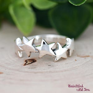 May include: A silver ring featuring a repeating star design. The ring has a polished finish and is set against a wooden surface with a blurred green background. The ring's design includes five evenly spaced, three-dimensional stars.