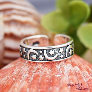 May include: A silver-colored ring with a celestial design featuring crescent moons, stars, and dots. The ring is set against a backdrop of a seashell and greenery. The ring's band is decorated with the celestial pattern.