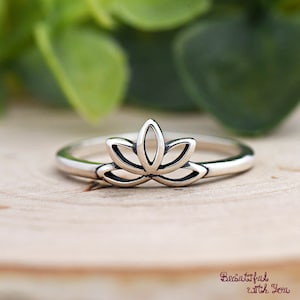 May include: A silver ring featuring a lotus flower design. The ring's band is thin, and the lotus flower is the focal point, with its petals clearly defined. The ring is set against a wooden surface with a blurred green background. The text "Beautiful with You" is visible.