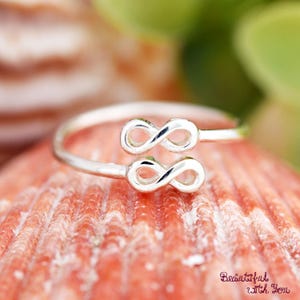 May include: A silver-colored ring featuring two infinity symbols. The ring is set against a blurred background of a pinkish-red surface and green foliage. The ring is a delicate piece of jewelry.