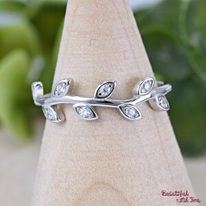 May include: A silver ring featuring a delicate leaf design. Each leaf is adorned with a small, sparkling gemstone. The ring is displayed on a wooden stand, showcasing its intricate details and elegant style.