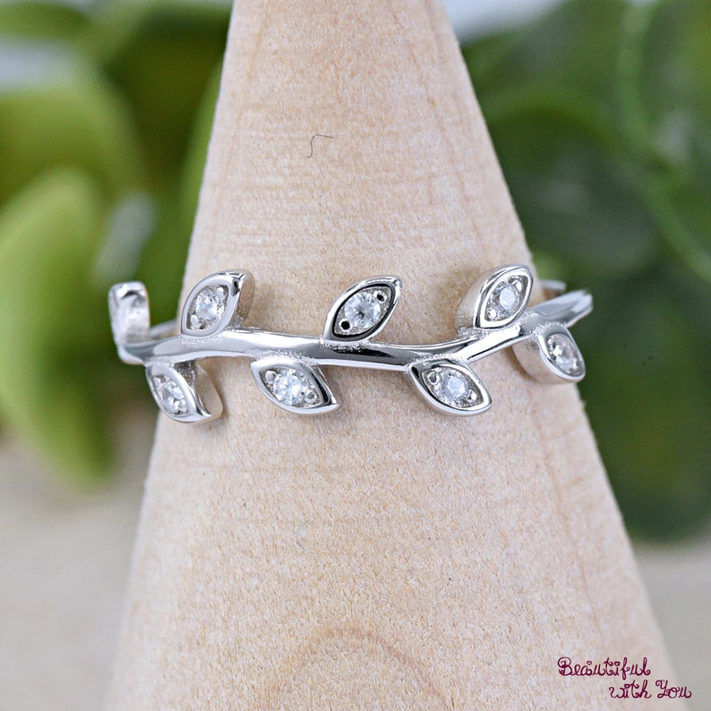 May include: A silver ring featuring a delicate leaf design. Each leaf is adorned with a small, sparkling gemstone. The ring is displayed on a wooden stand, showcasing its intricate details and elegant style.