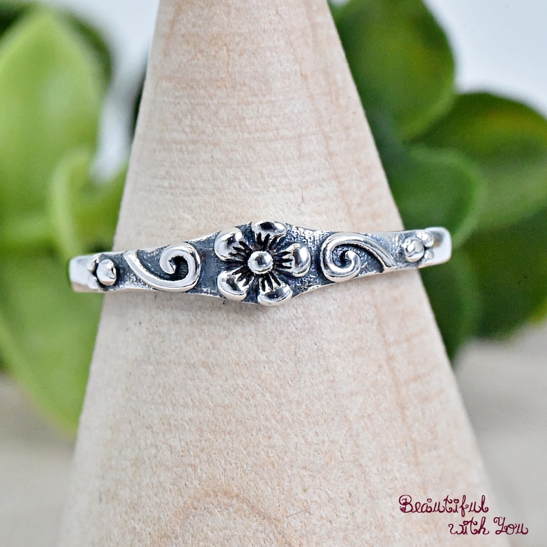 May include: A silver ring with a delicate floral design. The ring features a small flower with a swirl pattern on either side.