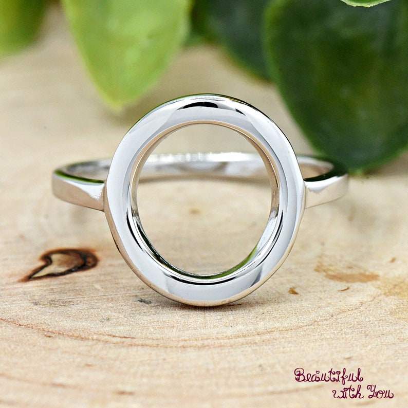 May include: A silver ring with a large, circular, open design. The ring is on a wooden surface.