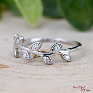 May include: A silver ring featuring a delicate leaf design. The ring has small, clear gemstones set in the leaves. The ring is on a wooden surface with a blurred green background. The ring is a piece of jewelry.