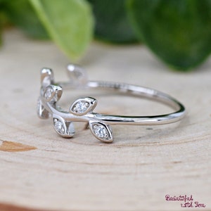 May include: A silver ring featuring a delicate leaf design. The ring has small, sparkling gemstones set into the leaves. The ring is displayed on a light-colored wooden surface, with a blurred green background.