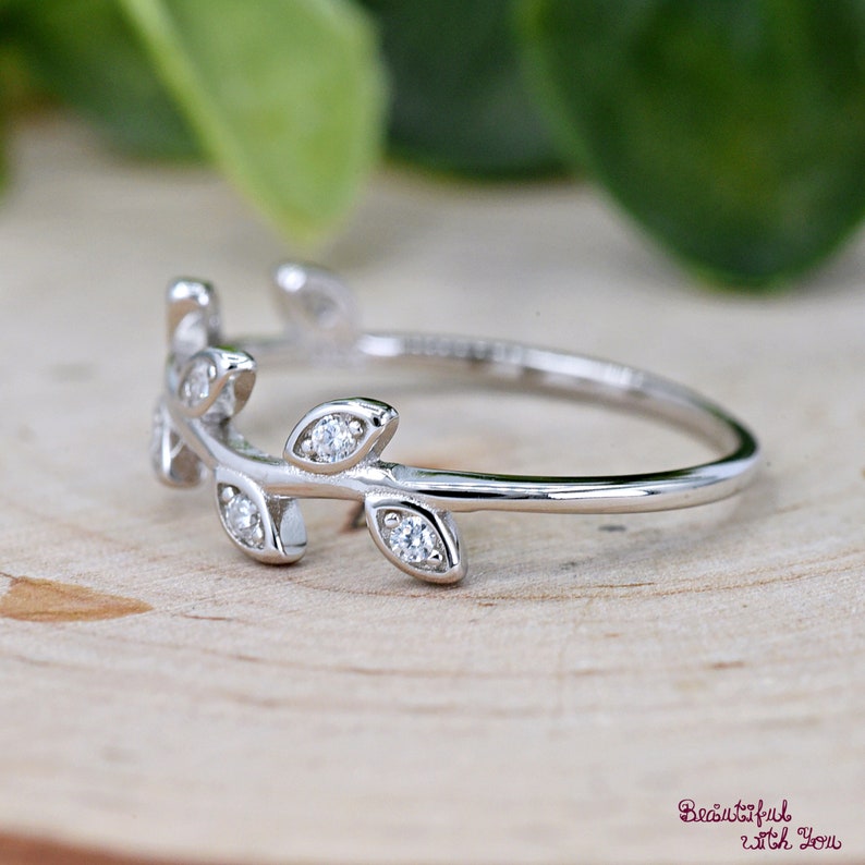May include: A silver ring featuring a delicate leaf design. The ring has small, sparkling gemstones set into the leaves. The ring is displayed on a light-colored wooden surface, with a blurred green background.