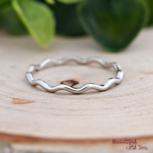 May include: A silver-colored ring with a wavy, undulating design. The ring is set against a wooden surface with green foliage in the background. The ring's design is a continuous wave pattern.