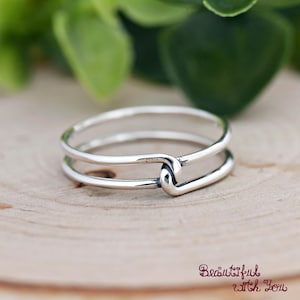May include: A silver-colored ring with a double-band design, featuring a knot-like detail at the center. The ring is set against a wooden surface with a blurred green background. The words "Beautiful with You" are written in cursive.